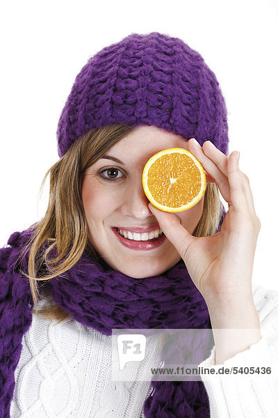 Young woman wearing winter clothes  holding an orange in front of her eye