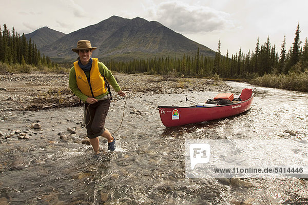 Young woman lining  wading  pushing  pulling a canoe in shallow water  Wind River  Peel Watershed  Northern Mackenzie Mountains behind  Yukon Territory  Canada