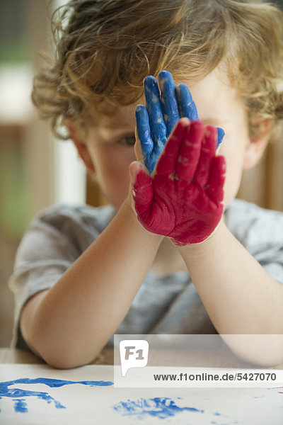 Kleiner Junge  der mit Farbe bedeckte Hände vor seinem Gesicht hält.
