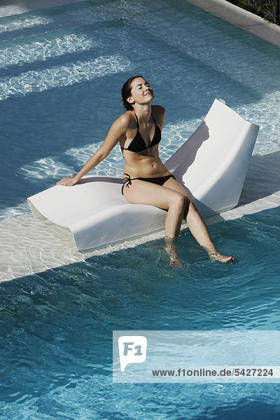 Junge Frau sitzt auf einem Liegestuhl am Pool und baumelt mit den Füßen ins Wasser.