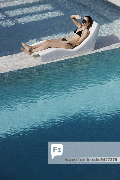 Young woman reclining on poolside deckchair  shading eyes with hand