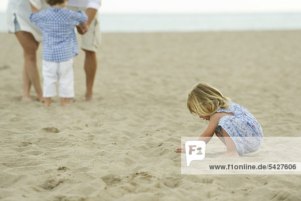 Kleines Mädchen spielt im Sand am Strand  Familie im Hintergrund
