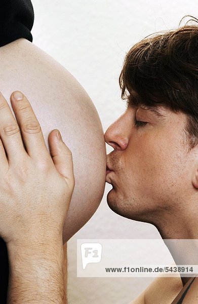 Paar  schwangere Frau  ihr Bauch wird von ihrem Mann vor weißem Hintergrund geküsst.