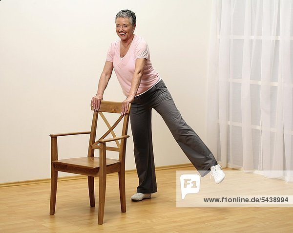 ältere Frau macht Gymnastik mit Stuhl - Bein seitlich heben - Muskelkraft - Seniorin