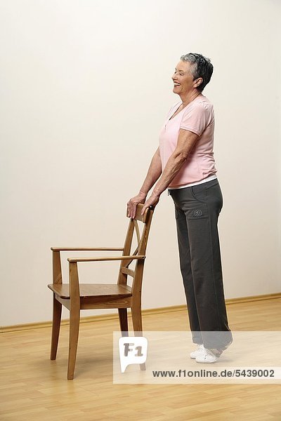 older woman doing gymnastics with a chair - woman is standing on tiptoe - pumping - muscularity - senior