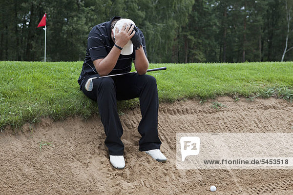Ein Golfer sitzt am Rande einer Sandfalle  Kopf in den Händen.
