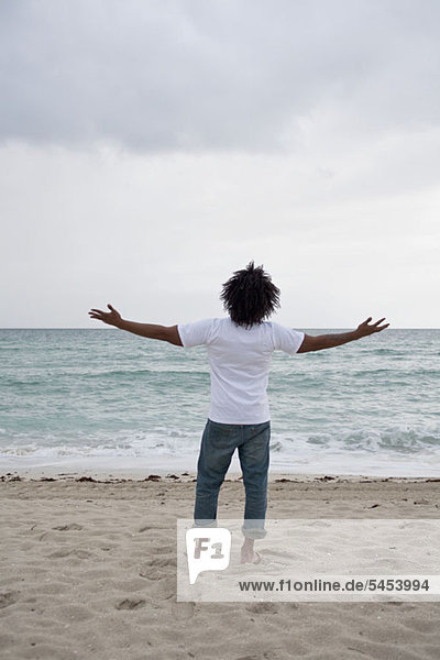A young man standing on beach with arms outstretched  rear view