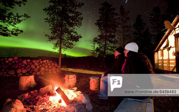 Junges Paar  Frau und Mann sitzen am Lagerfeuer und beobachten Nordlichter  Polarlichter oder Aurora Borealis  grün  erleuchtetes Zelt  Hütte  bei Whitehorse  Yukon Territory  Kanada