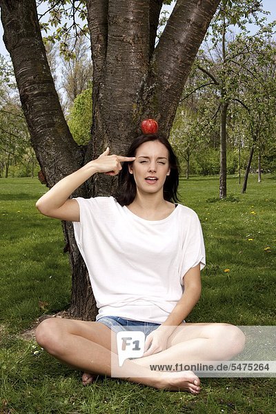 Junge Frau sitzt mit einem Apfel auf dem Kopf auf einer Wiese