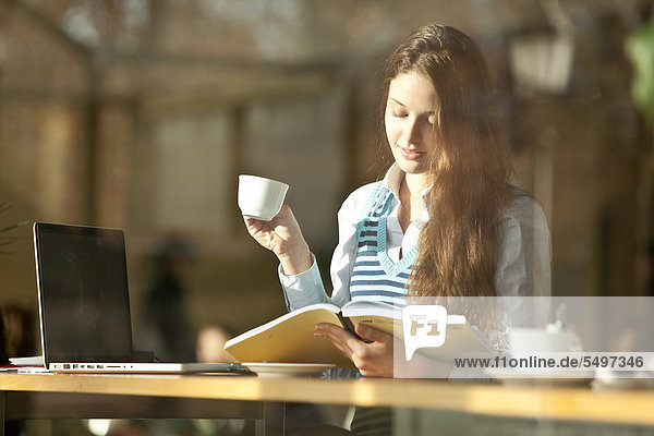 Studentin mit Laptop und Lektüre sitzt in Cafe  durch Fensterscheibe gesehen