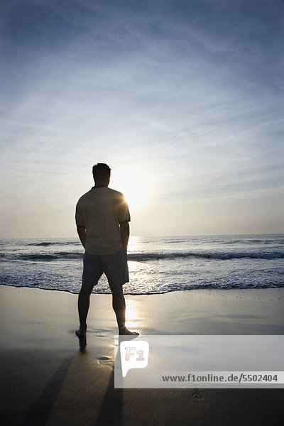 Caucasian Mid Adult Man Standing Alone On Beach Looking At Ocean At Sunrise