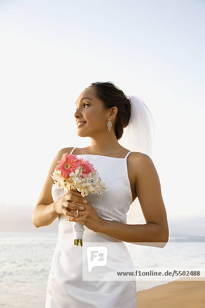 Young adult female Caucasian bride holding bouquet on beach.