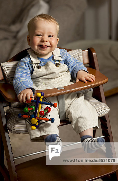 Kleiner Junge  8 Monate  mit Latzhose und Spielzeug im Kinderstuhl  streckt die Zunge raus  Deutschland
