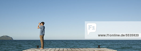 Mann am Pier stehend mit Blick auf das Meer durch ein Fernglas  Seitenansicht