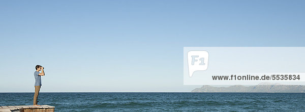 Mann in der Ferne steht auf dem Pier und schaut durch ein Fernglas auf das Meer.