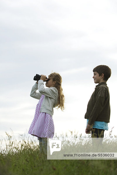 Girl standing in field with boy  looking through binoculars