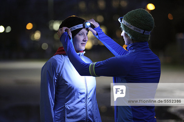 Zwei junge Frauen laufen in der Abenddämmerung im Winter  ziehen sich Reflektorbänder und LED-Stirnlampen an  mit reflektierender  wind- und wasserdichter Funktionsbekleidung