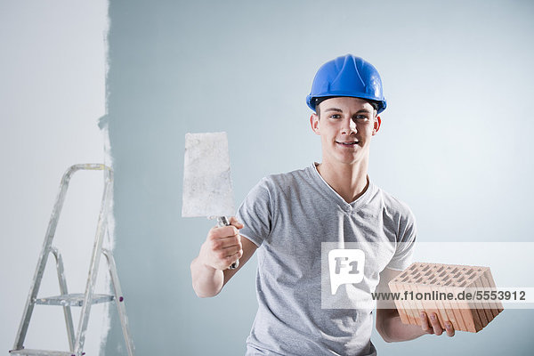 Young man wearing hard hat holding trowel and brick