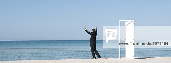 Geschäftsmann am Strand mit Blick auf den Ozean und erhobenen Armen,  Rückansicht