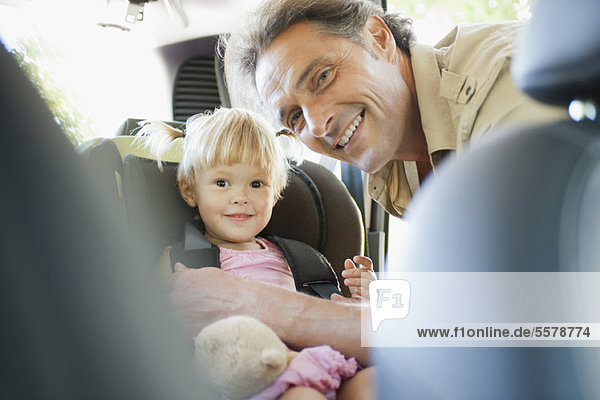 Vater befestigt kleines Mädchen im Autositz  beide lächelnd vor der Kamera