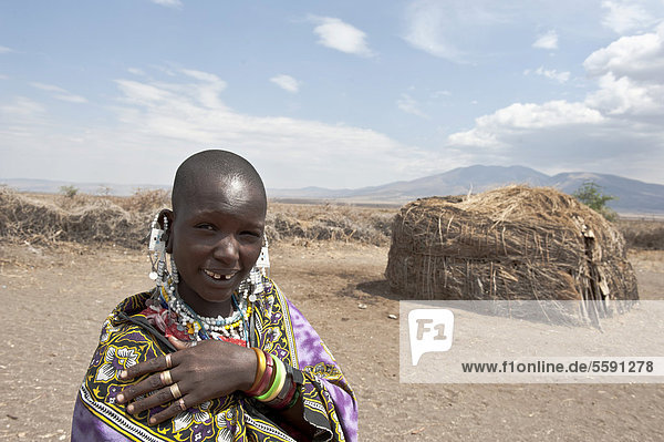 Portrait  Ethnologie  Frau der Massai trägt Schmuck  Maasai  kahl rasierter Kopf  vor Hütte aus Stroh und Ästen  Dorf Kiloki  Savanne  Ngorongoro Conservation Area  Serengeti Nationalpark  Tansania  Ostafrika  Afrika
