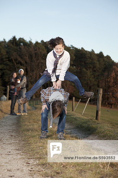 Germany  Bavaria  Children playing leapfrog and parents standing in background