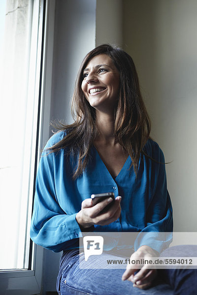 Mittlere erwachsene Frau am Fenster mit Handy  lächelnd