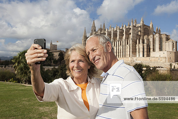 Spanien  Mallorca  Palma  Seniorenpaar lächelnd beim Fotografieren mit der Kathedrale Santa Maria  Portrait