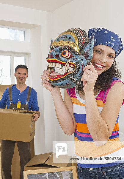 Frau hält asiatische Maske mit Mann im Hintergrund  lächelnd  Portrait