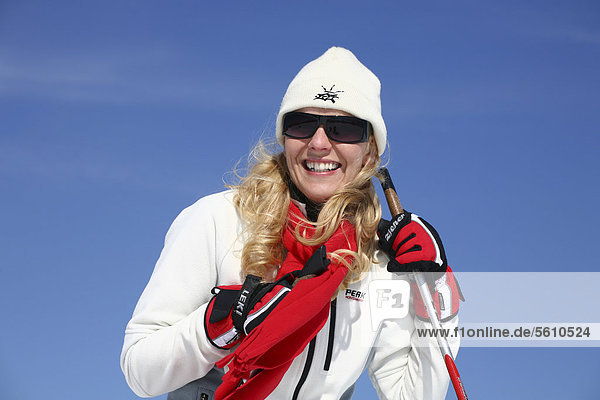 Young woman  about 25 years  wearing a bright jacket  a cap  dark sunglasses and a red scarf  against a blue sky  holding a ski stick  smiling  Thuringian Forest mountains  Thuringia  Germany  Europe