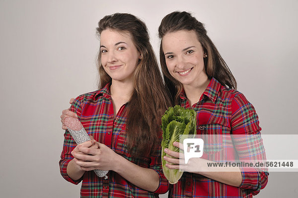 Zwillingsschwestern  die eine mit einer Salami in der Hand  die andere mit einem Salatkopf