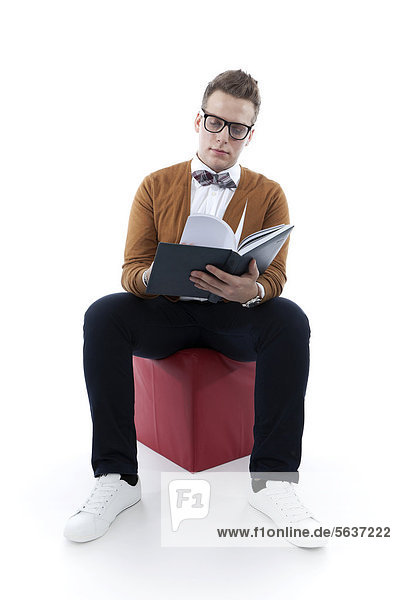 Junger Mann mit Brille und Fliege blättert in Buch  Agenda und sitzt auf rotem Sitzwürfel