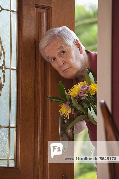 Ein älterer Mann  der durch eine offene Tür mit einem Blumenstrauß schaut.