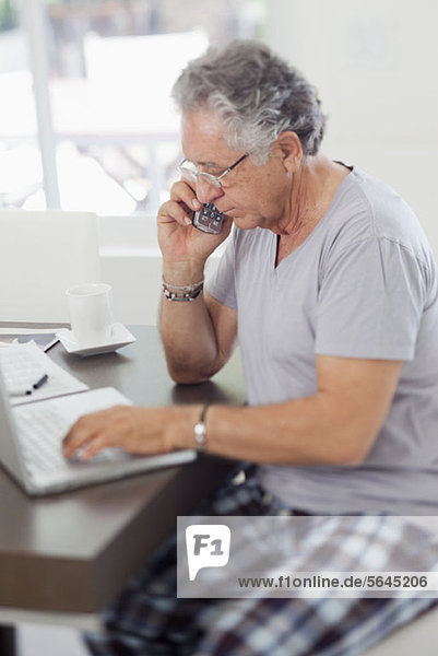 Ein älterer Mann  der einen Laptop benutzt und zu Hause mit einem schnurlosen Telefon spricht.