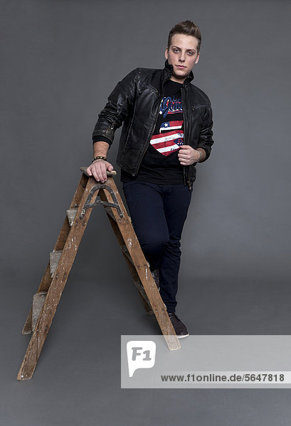 Junger Mann mit Lederjacke und Jeanshose stützt sich auf alte Holzleiter