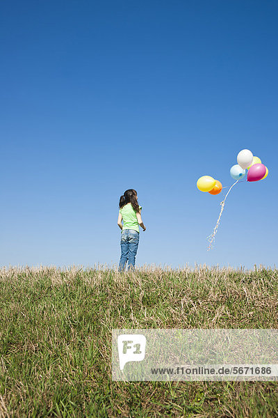 Mädchen steht auf einer Wiese und schaut auf Luftballons