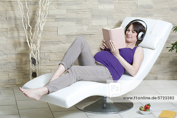 Germany  North Rhine Westphalia  Pregnant woman reading book and listening music