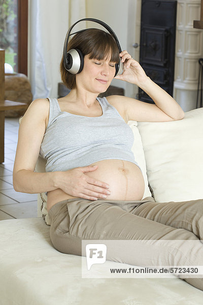 Deutschland  Nordrhein-Westfalen  Schwangere mit Kopfhörer und Sitzen auf Couch