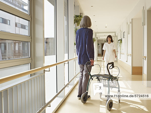 Senior women holding walking frame in corridor  caretaker in background