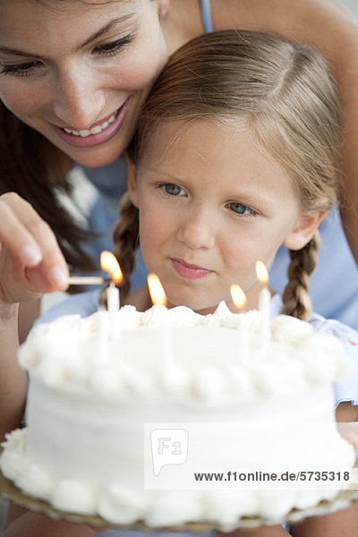 Mädchen sieht zu  wie ihre Mutter Kerzen auf dem Geburtstagskuchen anzündet.
