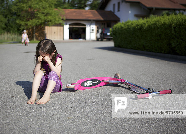 Dreijähriges Mädchen ist mit Roller hingefallen und sitzt weinend auf dem Boden