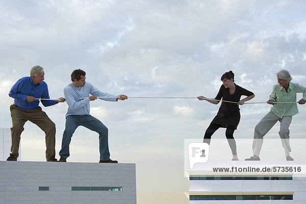 Überdimensionale Männer und Frauen stehen auf Dächern und spielen Tauziehen.