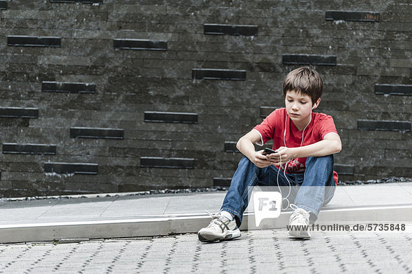Junge sitzt auf dem Bürgersteig und hört Musik mit Kopfhörern.