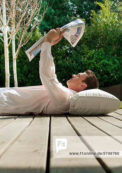 Businessman lying on decking reading newspaper