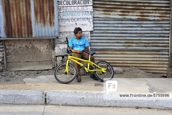 Jugendlicher mit gelbem Fahrrad sitzt gelangweilt vor einer Wellblechhütte  Armenviertel Lomas de Santa Faz  Guatemala Stadt  Guatemala  Mittelamerika