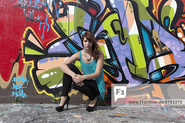 Junge Frau mit türkisem Top  schwarzen Leggins und hohen Schuhen vor Graffitiwand