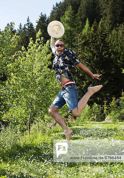 Laughing man wearing sunglasses and a Hawaiian shirt with a straw hat  jumping in a flowering meadow