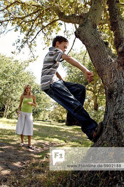 Side profile of a boy climbing a tree with his mother standing in a park