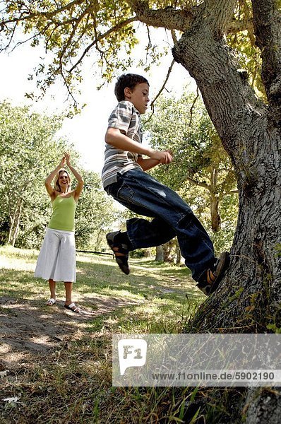 Side profile of a boy climbing a tree with his mother clapping in a park
