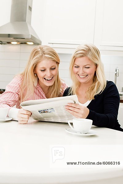 Two young women reading newspaper in kitchen